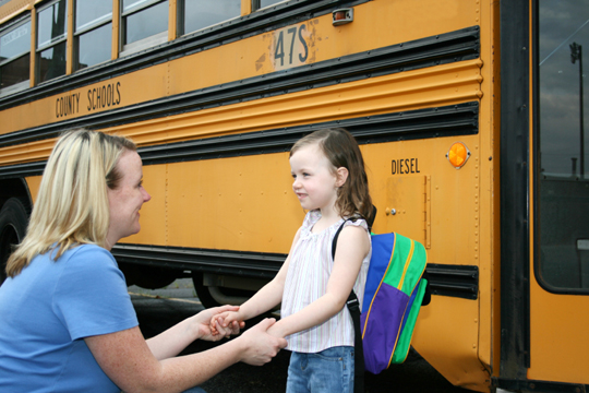 Hero_HomepageSlider_BackToSchool Image of a woman lovingly holding a small girl's hands in front of a school bus